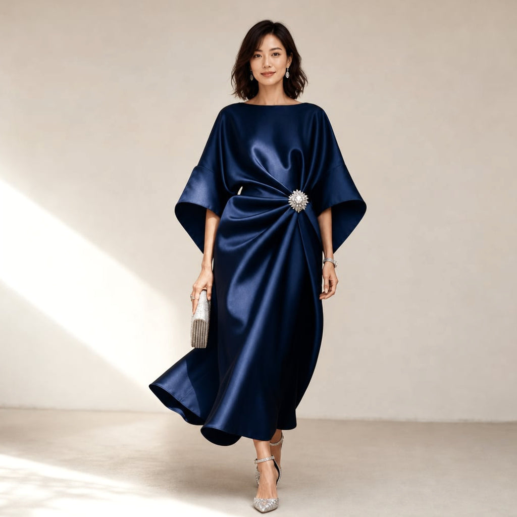 Modelo lleva vestido midi azul marino de punto con acabado satinado, mangas kimono y broche de cristal, posando de frente