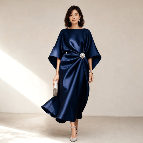 Modelo lleva vestido midi azul marino de punto con acabado satinado, mangas kimono y broche de cristal, posando de frente