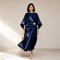 Modelo lleva vestido midi azul marino de punto con acabado satinado, mangas kimono y broche de cristal, posando de frente