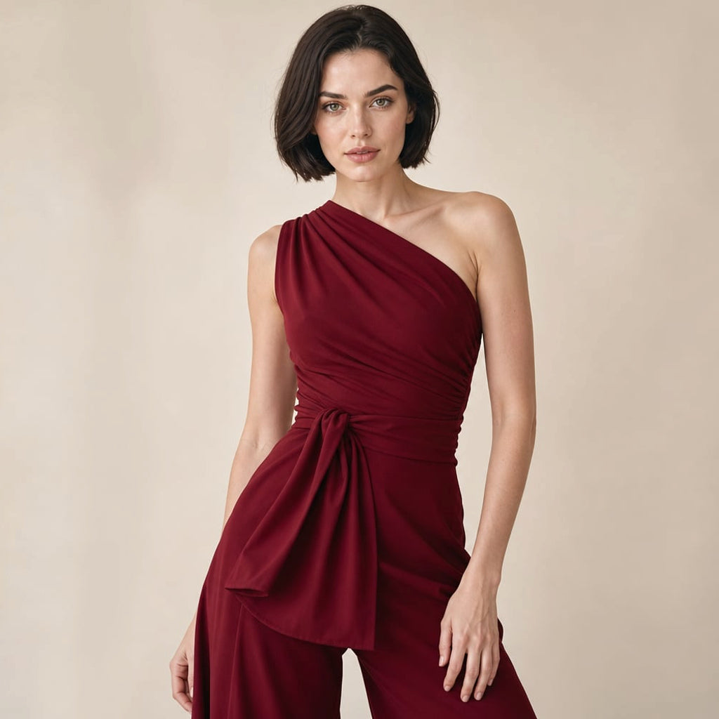 Modelo de pie usando jumpsuit borgoña de un solo hombro con pierna ancha y moño en la cintura natural