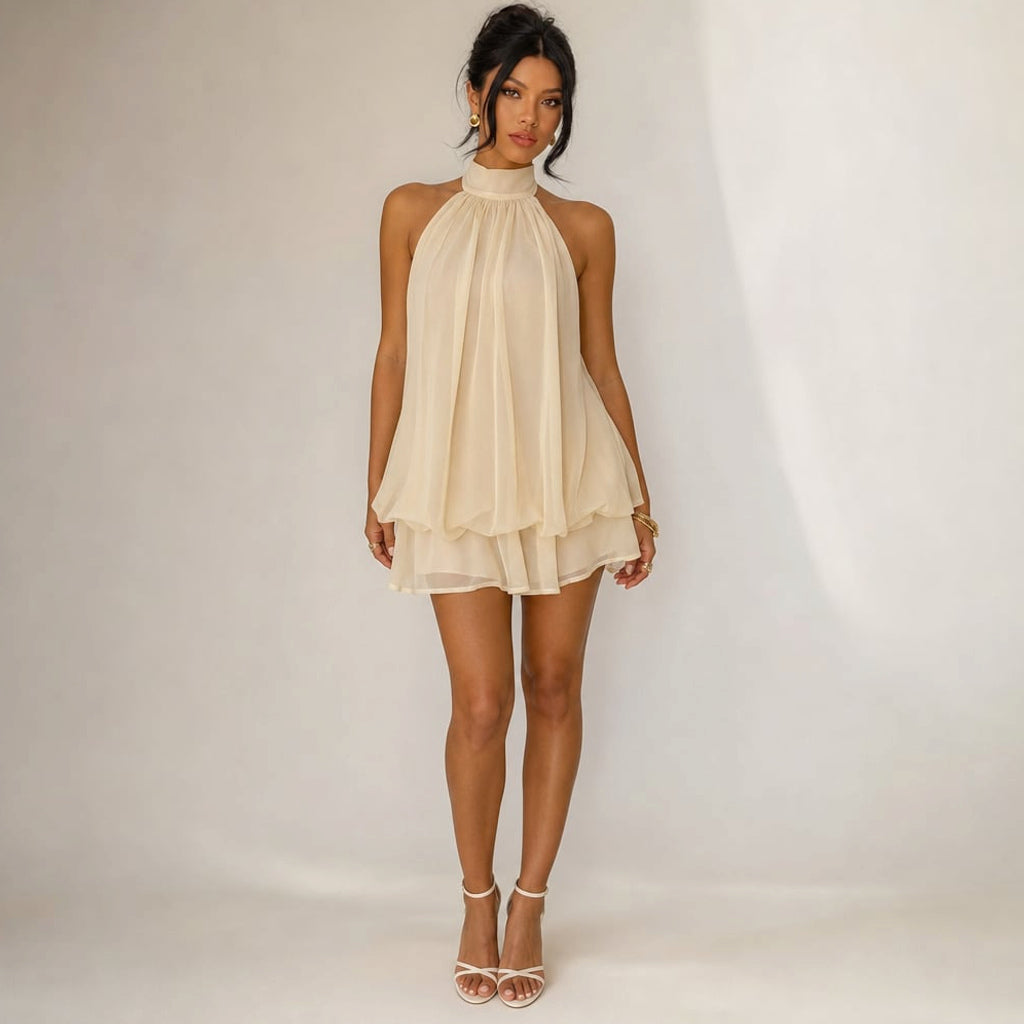 Vestido mini de gasa color beige con cuello halter anudado, holanes escalonados en la falda y espalda abierta, sobre fondo neutro